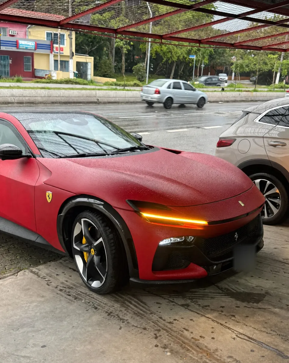 Ferrari Purosangue vermelha fosca com o farol aceso, na entrada da RM em dia de chuva